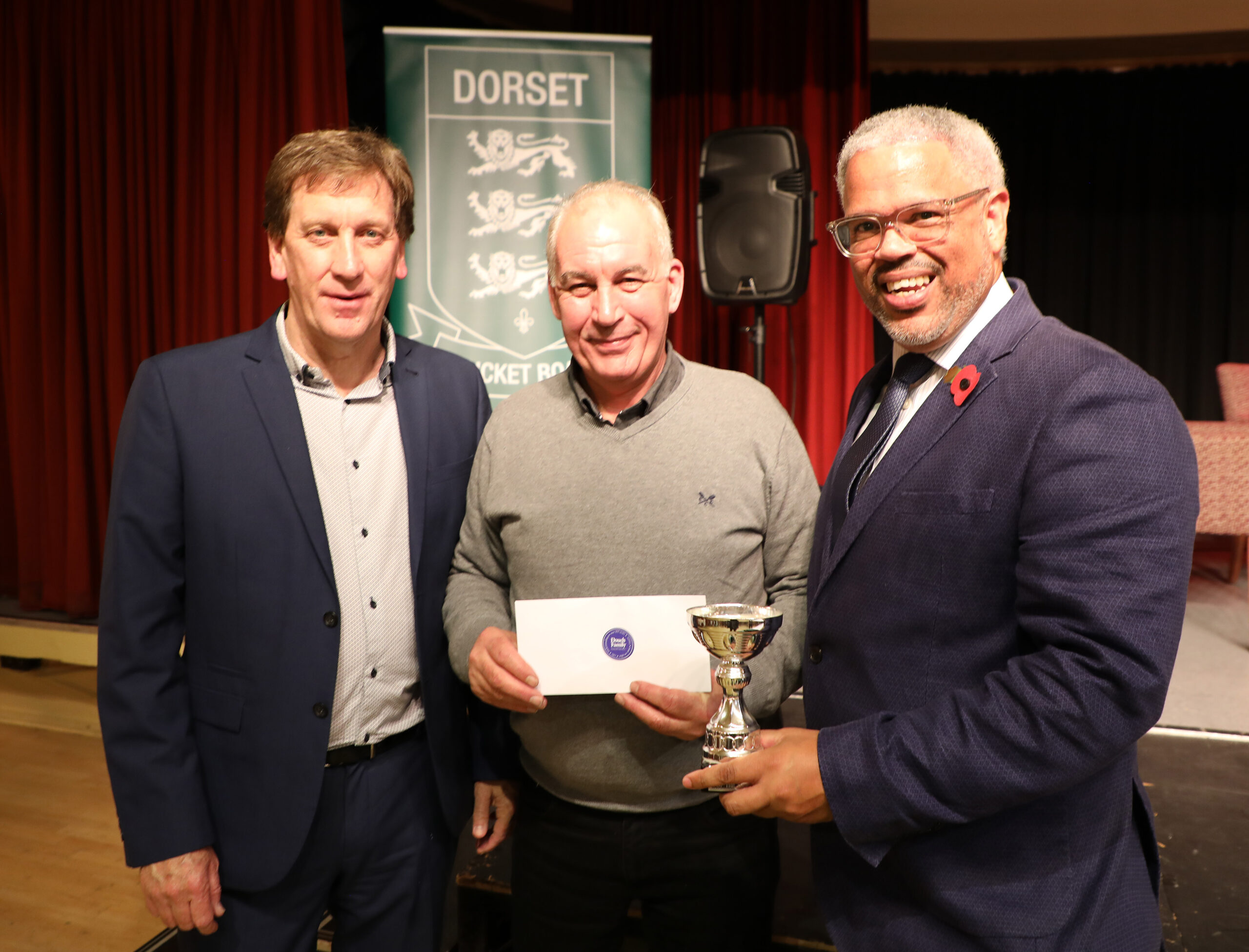 Three men smiling at the camera during an award presentation event. On the left, Kevan James, a former Hampshire CCC player, dressed in a blue blazer and casual shirt. In the center, Dave Abbott, Chairman of Blandford CC, holds an envelope and wears a grey sweater. On the right, Nick Douch from Douch Family Funeral Directors, stands with a silver trophy, wearing a smart blue suit and a red poppy pin. A banner for the Dorset Cricket Board is visible in the background.