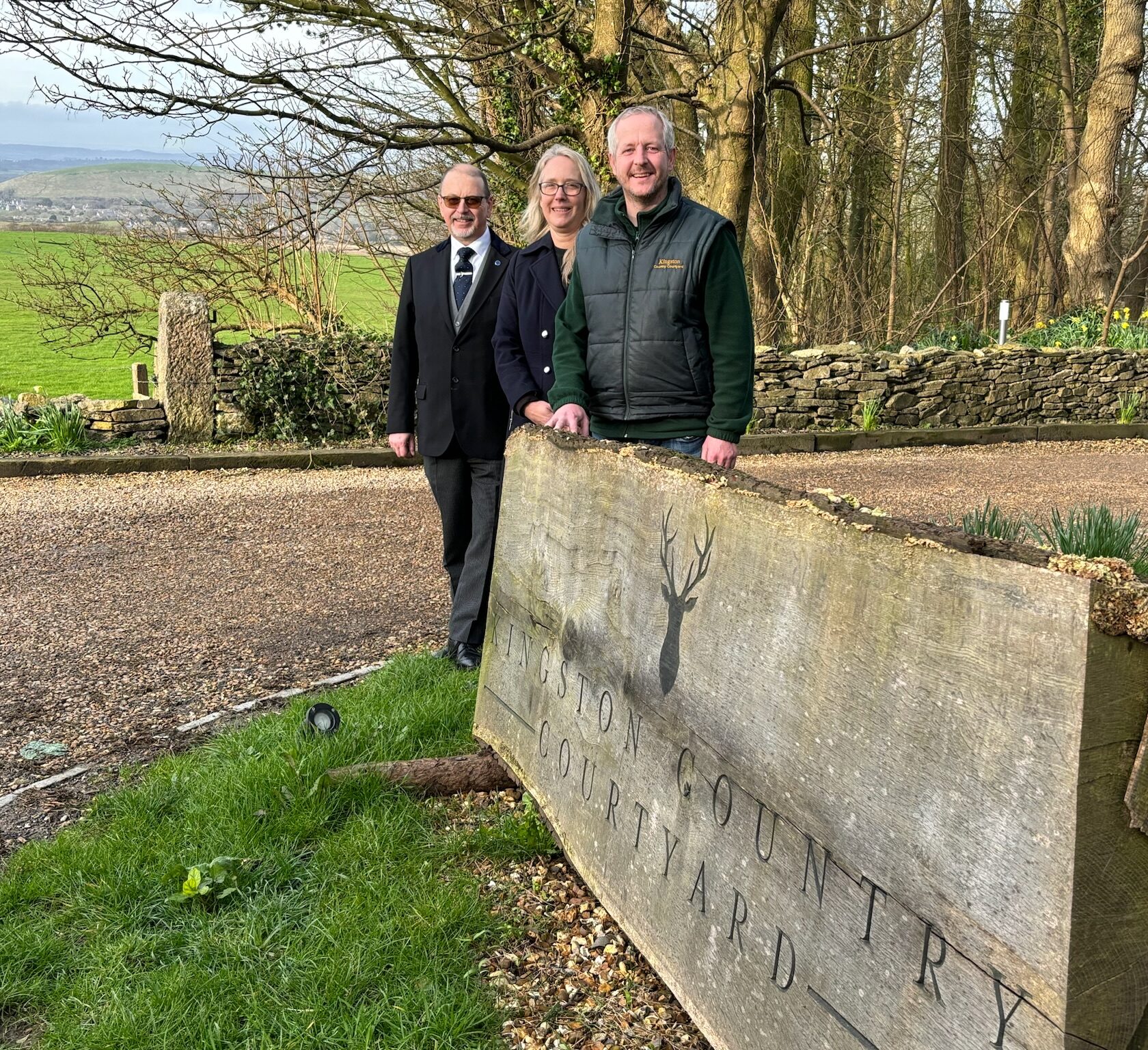 Three people standing next to a wooden sign at Kingston Country Courtyard with the lush Purbeck Hills in the background.