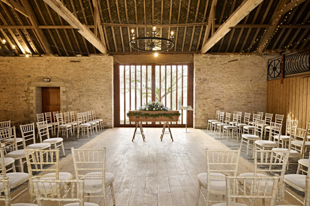 Interior view of Kingston Country Courtyard's rustic barn venue set up for a funeral service with white chairs and a casket covered with flowers at the forefront, with large windows revealing natural light.