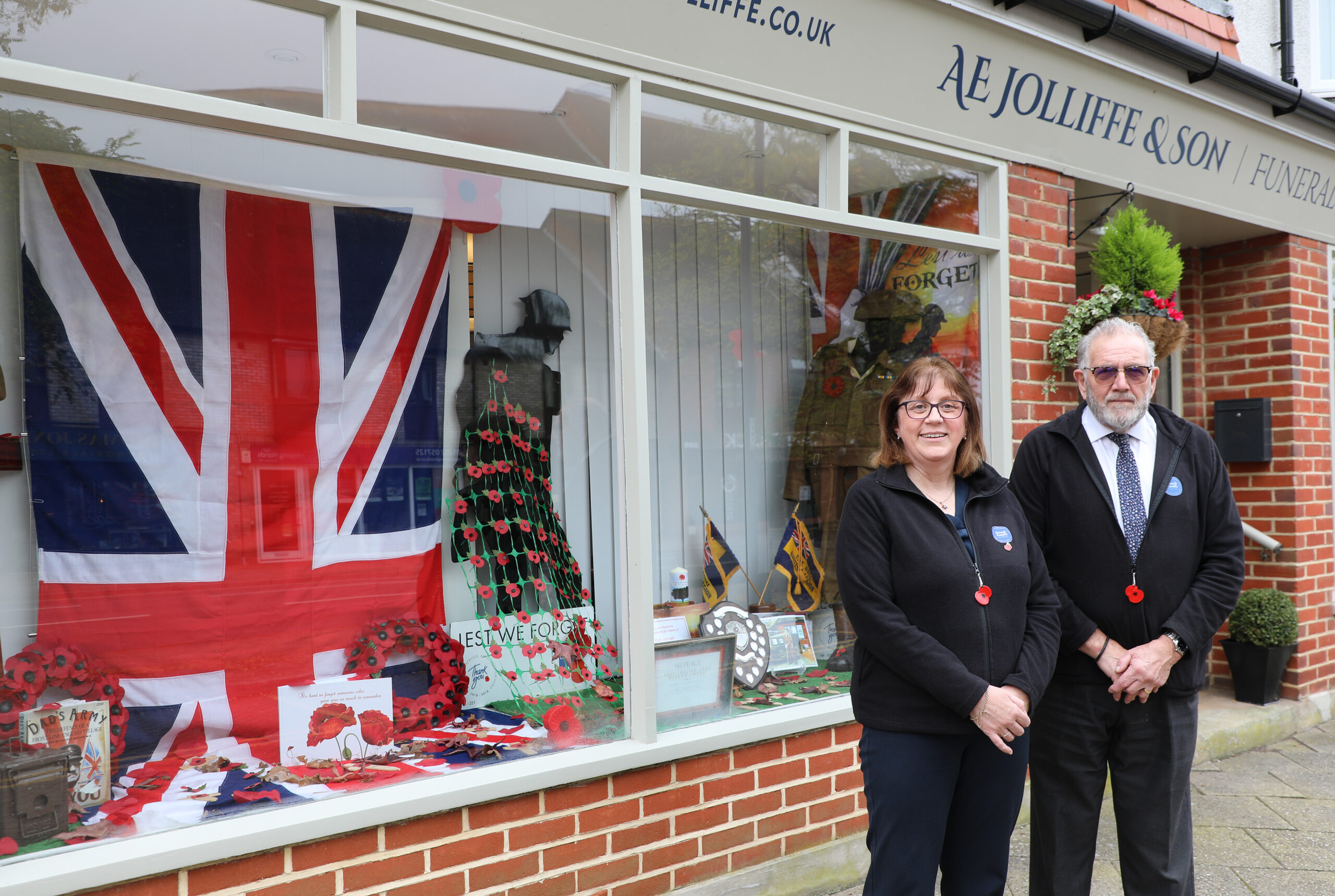 A man and woman stand outside AE Jolliffe & Son Funeral Directors, posing beside a storefront display dedicated to Remembrance Day. The window features a large Union Jack flag, a mannequin adorned with poppies, and various memorabilia with the words "Lest We Forget." Both individuals wear red poppy pins, symbolizing remembrance for those who served in the military.