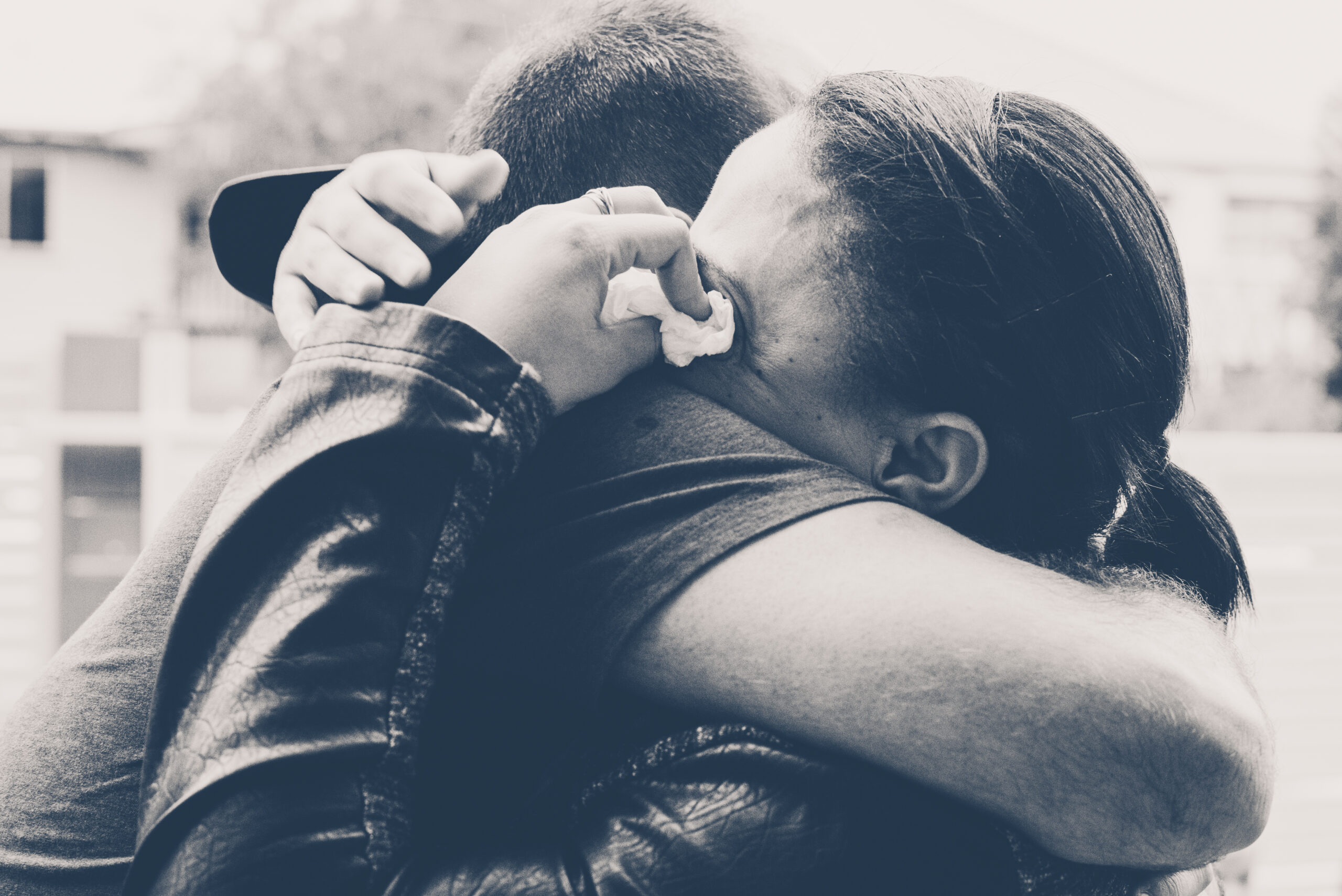 A grieving woman tightly embraces a man while crying into his shoulder, holding a crumpled tissue in her hand. The emotional moment captures the comfort of a supportive hug during a time of loss.